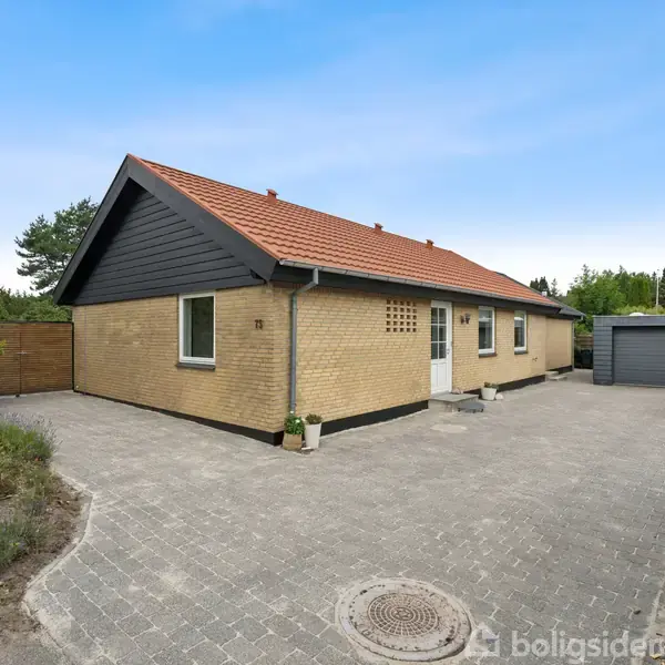 Et gult murstenshus med rødt tag står i en have omgivet af grønne træer. En flisebelagt indkørsel leder til en enkelt garage ved siden af huset. En postkasse er nær indkørslen.