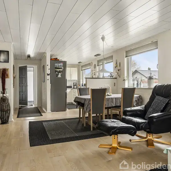 Stue indrettet med spisebord og fire stole ved vinduerne. En lænestol med fodskammel står ved siden af. Trægulve og lyst loft; køkkenet ses i baggrunden.