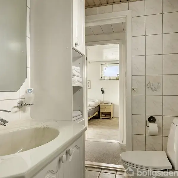 Badeværelse med hvidt toilet ved siden af et hvidt vaskeskab med spejl. Hylderne rummer håndklæder. En åben dør viser et soveværelse med seng og natbord i baggrunden.