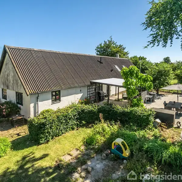 Et hvidt hus med mørkt tag står i haven. Der er en terrasse med borde, stole, og en parasol. Græs og buske omgiver huset. Solrig himmel i baggrunden.