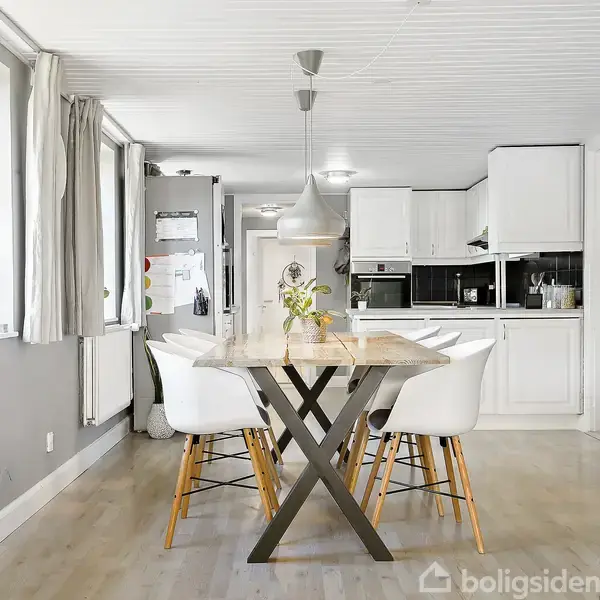 Et spisebord med seks stole står i midten af et lyst køkken-alrum. Bordet har en vase med blomster. Køkkenet har hvide skabe og sorte apparater i baggrunden. Gardiner dækker vinduerne.