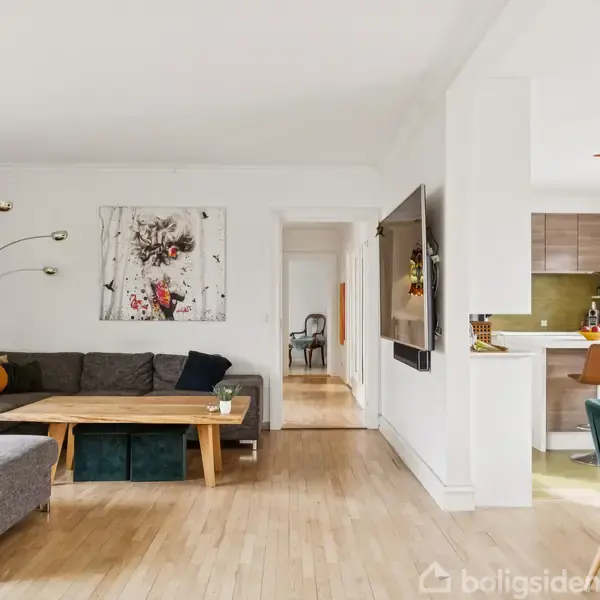 Grå sofa med puder står i stue, omkring et træbord, under en dekorativ lampe. Ved siden af er et moderne køkken med barstole. Lyse trægulve og kunst på væggen.