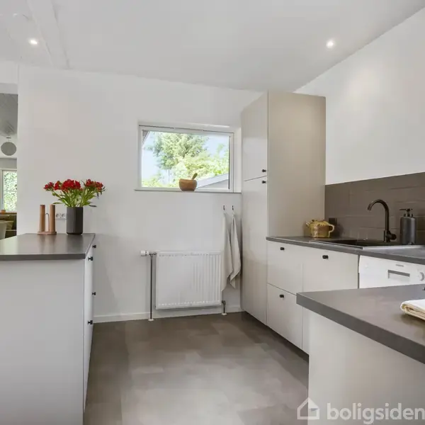 Et moderne køkken har hvide skabe, sort bordplade og en vask. En vase med røde blomster står ved et vindue, omgivet af et lyst rum med udsigt til en grøn have.