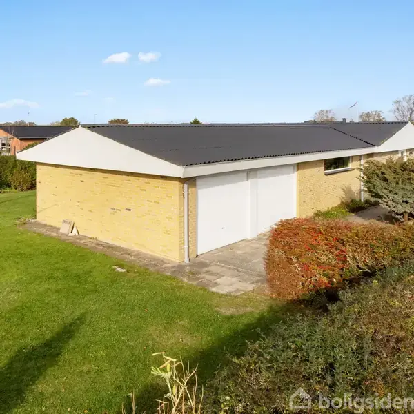 En gulstensbygning står stille i en have med græsplæne og buske omkring. Bygningen har et sort tag og en garageport. Himlen er blå med få skyer.