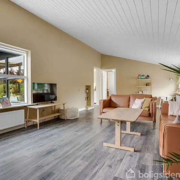 Stue med lysebrune vægge og hvide loftpaneler. To sofaer omgiver et sofabord. Der er en reol med dekorationer, en stor vindue med planter og et fjernsyn.