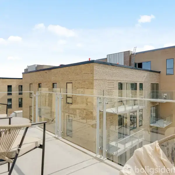 En moderne altan med glasrækværk tilbyder udsigt over et boligkompleks af lyse murstensbygninger. En rattan stol og plante er placeret på altanen under en klar himmel.