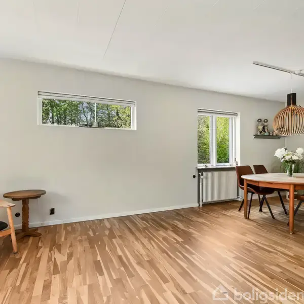 Giraffefigur står ved siden af et lille rundt bord, mens et spisebord med blomsterdekorationer er placeret nær vinduerne. Trægulvet strækker sig gennem det lyse rum.