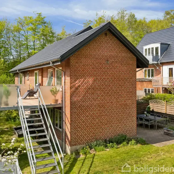 Rødt murstenshus med en stor terrasse til venstre, hvor der står stole. En udendørs trappe fører op til terrassen. Huset står i et grønt, beplantet område med høje træer.