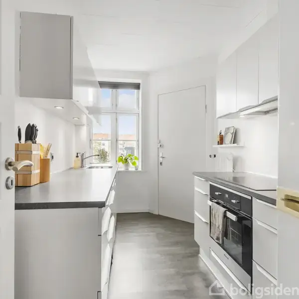 Et moderne køkken har hvide skabe og en mørk bordplade. En kogeplade og ovn er integreret. Et vindue med planter skaber lys og rummet føles rummeligt og åbent.