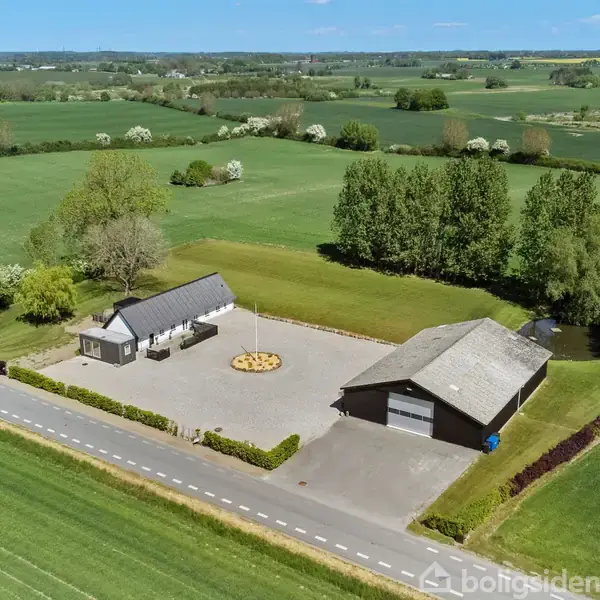 Et hus og en stor garage står på en åben gårdsplads omgivet af grønne marker og træer. En asfaltvej løber langs ejendommen under en blå himmel.