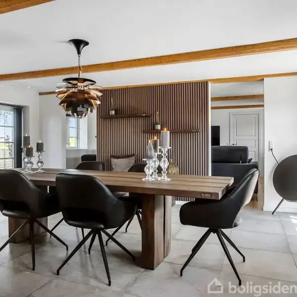 Spisebord i træ med seks sorte stole omgivet af lys, moderne stue med hvide vægge og træloftsbjælker. En stilfuld lampe hænger over bordet, og der er et møbel med stearinlys i baggrunden.