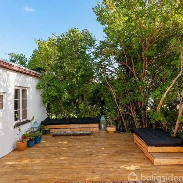 Terrasse med træmøbler står på en trædæk i en have. Hvide vægge og flere potteplanter omkranser området. Baggrunden domineres af grønne træer under en klar blå himmel.