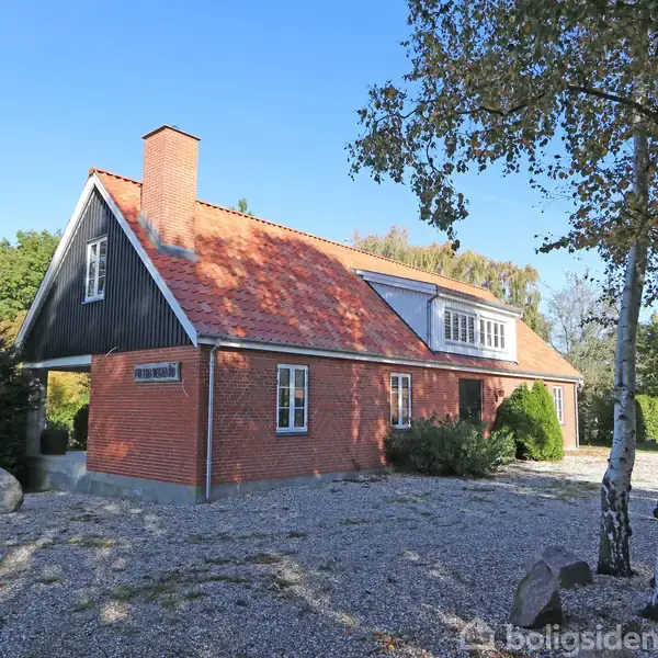 Et rødt murstenshus med rødt tegltag står i en grusbelagt have med træer. På ydervæggen hænger et skilt med teksten "OERNSTED".