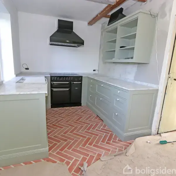 Et køkken med grå skabe, et sort komfur og en emhætte. Rødlig sildebensmønstret gulv. Omgivelserne inkluderer et lyst rum under renovering med ubehandlede vægge og kabler.