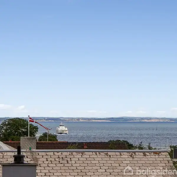 Et skib sejler på havet med klar himmel i baggrunden. Foran ses hustage med en dansk flagstang.