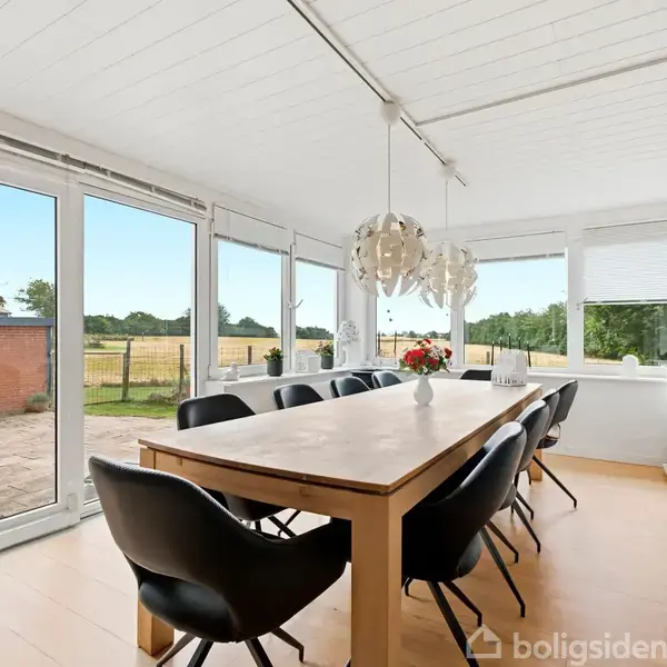 Et langt træbord omgivet af sorte stole står i et lyst spisestueområde. Store vinduer og glasdøre giver udsigt til en terrasse og grønne områder udenfor.