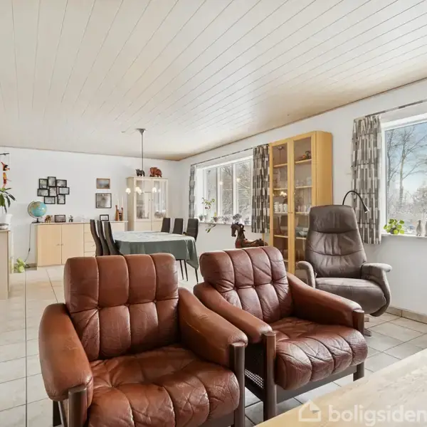 Brune lænestole står i en stue med lyse fliser. Et spisebord er i baggrunden ved siden af et vitrineskab. Store vinduer lader dagslys strømme ind.