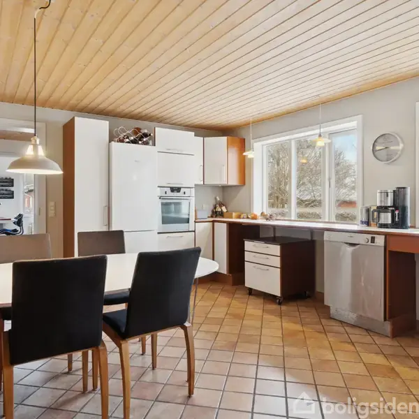 Et moderne køkken indrettet med et spisebord og fire stole under en hængende lampe. I baggrunden ses skabe, en ovn og store vinduer, der giver naturligt lys.