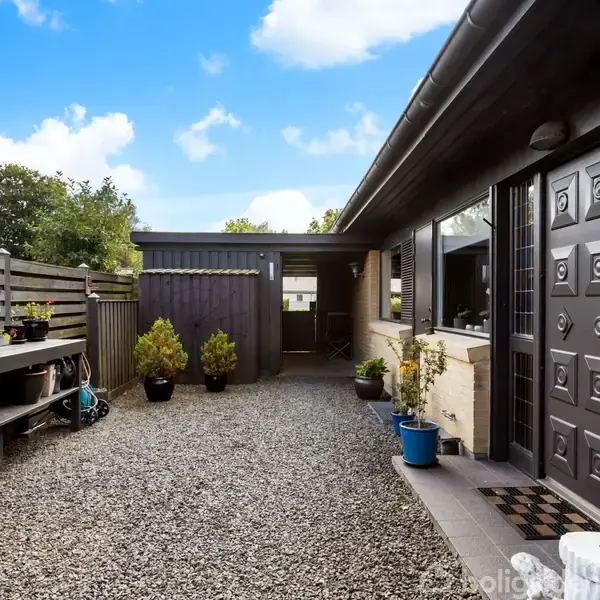 Et grusbelagt gårdhaveområde med en sortmalet bygning til højre, potteplanter langs væggen og en bænk til venstre. Solrigt vejr med nogle få skyer på himlen.