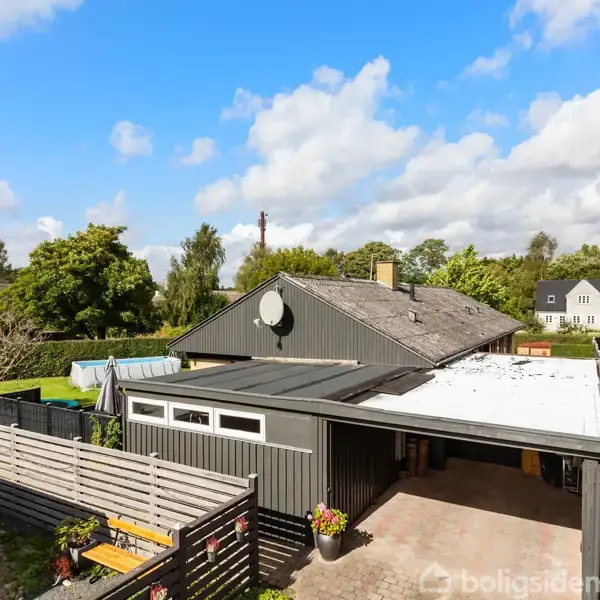 En grå etplansvilla med garage ligger i en have med træer og buske. Solen skinner, og himlen er blå med nogle skyer. Et par huse kan ses i baggrunden.
