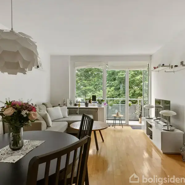 Rundt spisebord med blomster i vase står i en lys stue med sofahjørne, vinduer mod grøn udsigt, og lyst trægulv. Enkel indretning inkluderer reol og moderne lampe.