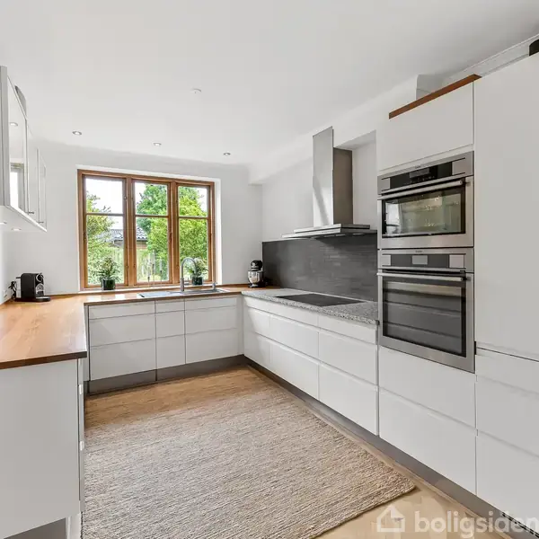 Et moderne køkken med hvide skabe og træbordplader. Der er en indbygget ovn og mikroovn. Store vinduer slipper naturligt lys ind og afslører en have udenfor.