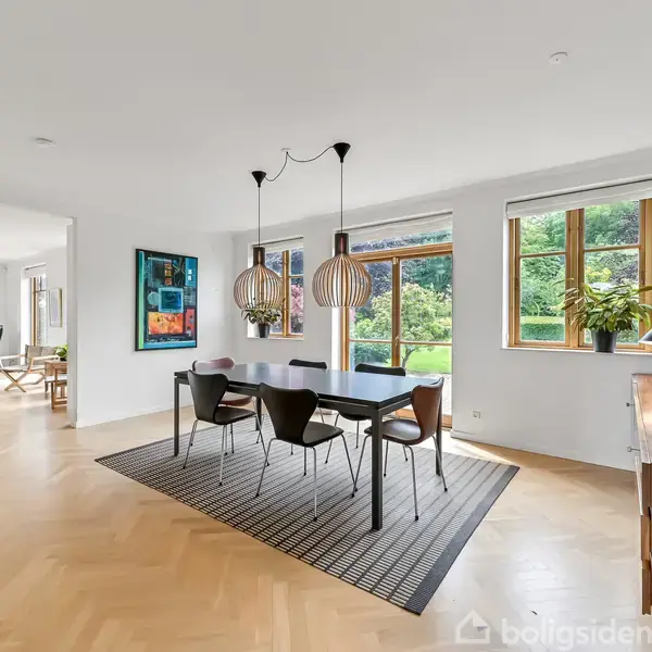 En spisestue med et langt, mørkt bord og fem stole står i et lyst rum. Store vinduer med udsigt til grønne omgivelser, dekoreret med moderne kunst på væggene.