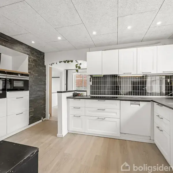 Køkken med hvide skabe og sorte klinker, indbygget ovn og mikrobølgeovn. Der er trælaminatgulv og vinduer, der giver naturligt lys. Moderne indretning med en minimalistisk stil.