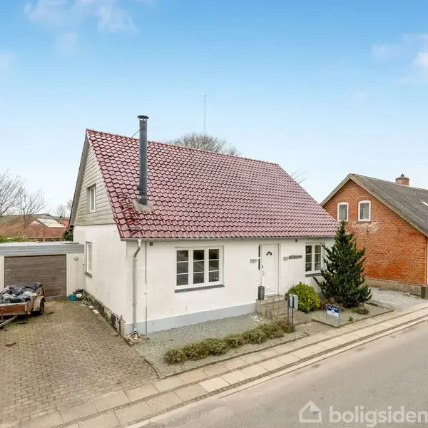 Hvidt hus med rød tagstenstag står ved vejkant. Lige op ad, et rødt murstenshus. Omgivet af brolagt indkørsel og garage i en forstadslignende omgivelse.