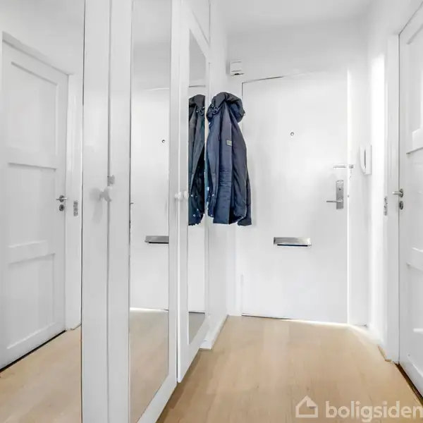 En entré med spejlskabe langs væggen, en jakke hængende ved døren, og lyse trægulve. To små billeder hænger på væggene.