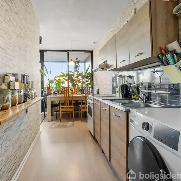 Et smalt køkken med over- og underskabe, en vaskemaskine og bordplade ved siden af en række krydderiglas. I baggrunden er et spiseområde med planter og stole.