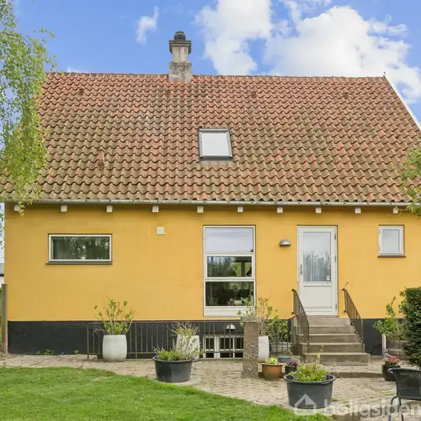 Et gult hus med rødt tegltag har trapper op til en hvid hoveddør. Foran er en have med græsplæne og krukker. Himlen er let skyet.
