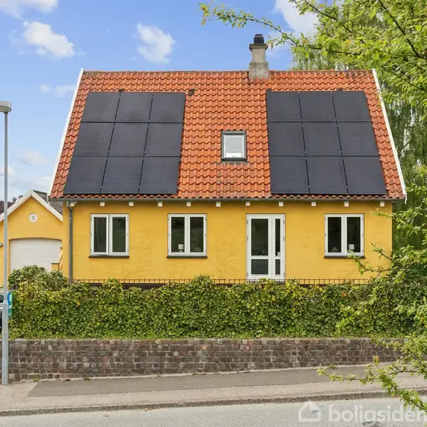 Gul hus med rødt tag og solpaneler, står i en hyggelig beboelse. Omkring er der hæk, træer og en belagt indkørsel med skraldespande foran en garage i baggrunden.