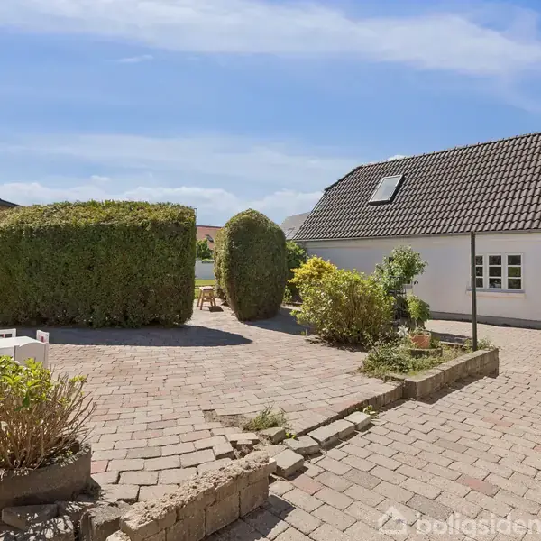 En brostensbelagt gårdhave med klippede buske og blomsterkrukker omkranser et hvidt hus med mørkt tag og ovenlysvinduer. Et træ- og græsbevokset område ses i baggrunden.