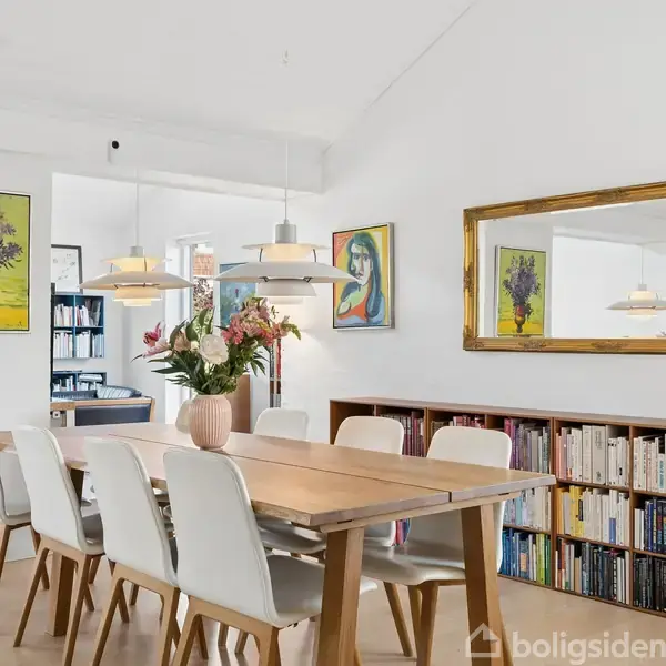 Spisebord med hvide stole er hovedfokus, dækket med en blomsterkande. Det står i en lys stue med kunstværker på væggene, en stor spejl og en bogreol i baggrunden.
