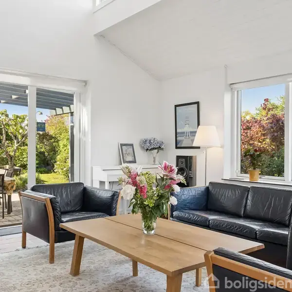 Sort læderhjørnesofa og træbord med blomster i en lys stue; store vinduer giver udsigt til frodig have med træer og terrasse, hvor et bord står dækket til fire.