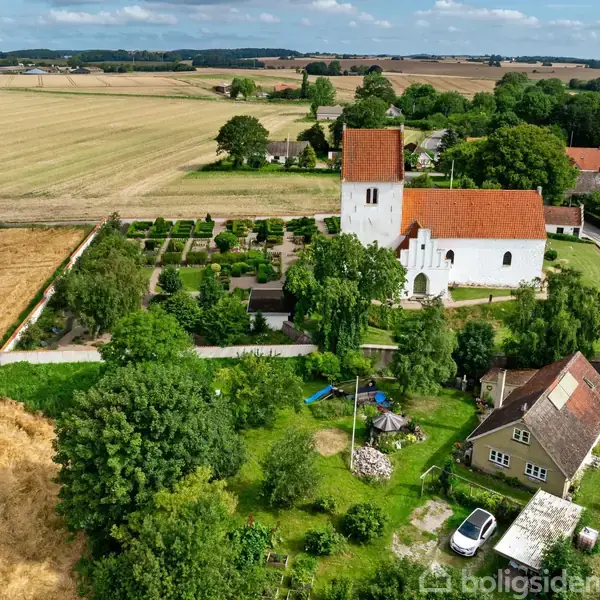 Hvid kirke med rødt tag står blandt grønne træer og marker. En mindre bygning og have ses i forgrunden, mens åbne marker strækker sig i baggrunden under en blå himmel.
