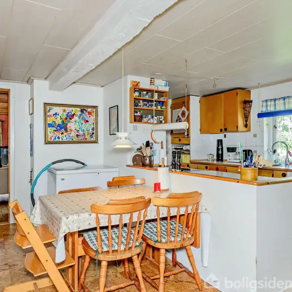 Et køkken-alrum med træstole om et bord med dug. Hylder og skabe fyldt med bøger og køkkengrej. Lys strømmer ind gennem et vindue med blå-ternet gardin.