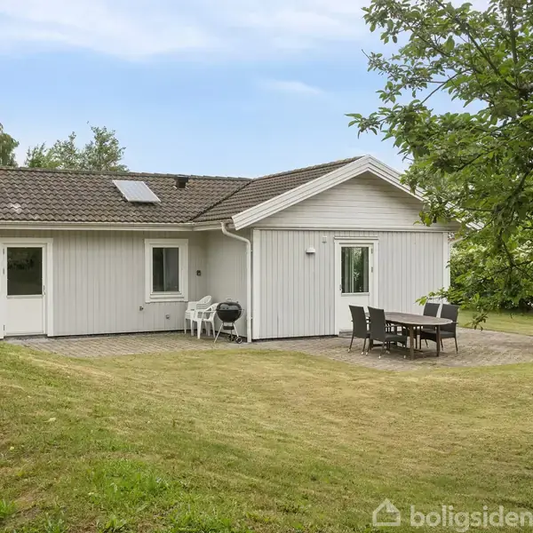 Hus set udefra med hvid facade og gråt tag, står på en grøn græsplæne. Terrasse med bord og stole ved siden af huset i en frodig have.