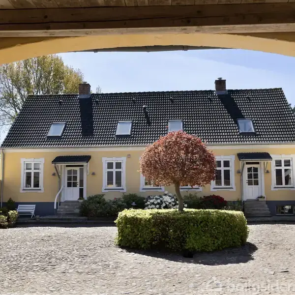 Et gult hus med mørkt tag står midt i en gårdsplads omgivet af træer og buske. To døre og flere vinduer vender ud mod pladsen under en blå himmel.