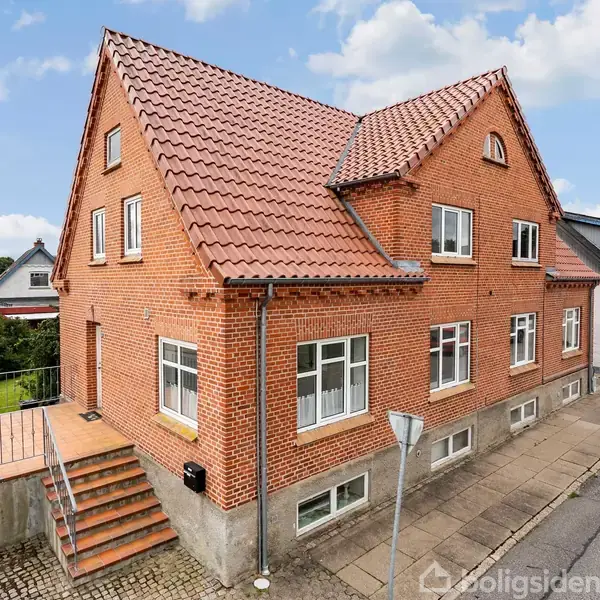 Rødt murstenshus med trekantet gavl og tegltag ligger på en fortovet gade. Trappe fører op til dør med gelænder. Omgivet af andre huse under en blå himmel med skyer.