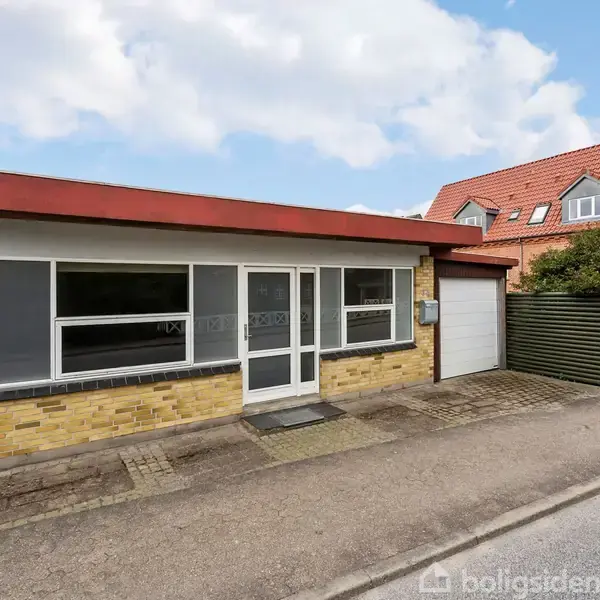 En enkeltstående bygning med gule mursten og fladt tag. Tre store vinduer og en glasdør vender ud mod en stille vej. Ved siden af ses et mørkegrønt plankeværk.