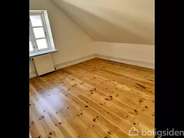 Et rum med et skråt hvidt loft og et vindue til venstre. Gulvet er lavet af lyst træ og spejler lys fra vinduet. Rummet er tomt og enkelt.