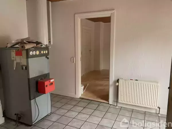 En kedel står stille i et flisebelagt rum, med en åben dør foran, der leder til et andet rum med trægulv. En radiator er monteret på væggen.