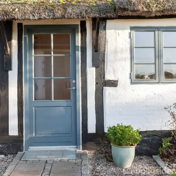 En blå dør indrammet af bindingsværk og vindue ved siden af, åbner ind til et hvidt hus med stråtag. Foran er en lille have med en grøn plante i en krukke.