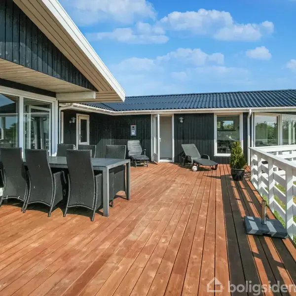 En stor træterrasse med sorte havemøbler står ved et moderne sortmalet hus med hvide detaljer. Solen skinner, og der er en åben udsigt over en græsplæne.