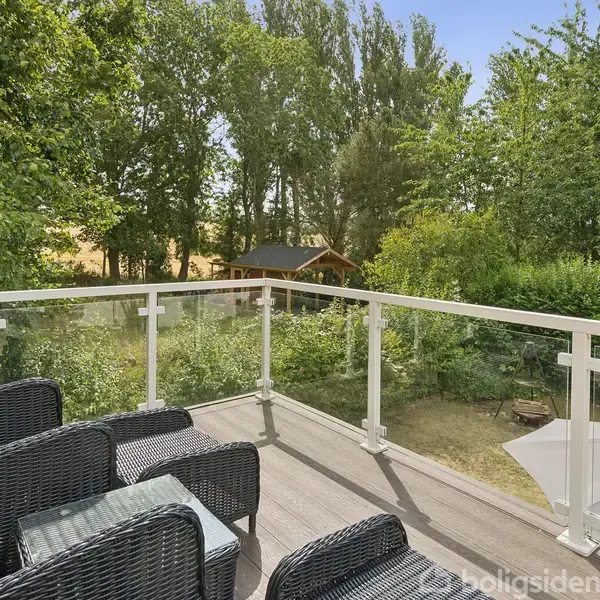 Terrasse med sorte fletmøbler placeret foran et glasrækværk; omgivet af frodig have med træer og buske, en parasol synlig i haven og en træbygning i baggrunden.