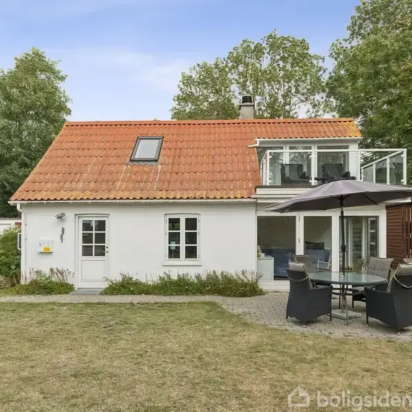 Huset har rødt tegltag og hvid facade, står i en have med græsplæne og træer, og foran er der havemøbler med en parasol.