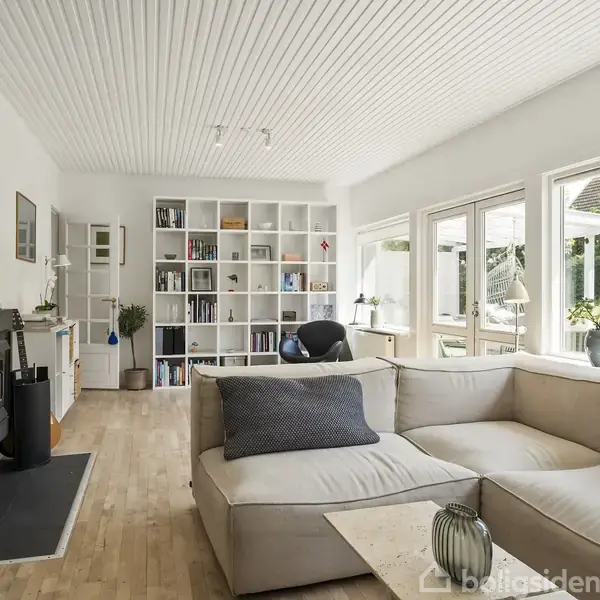 En stue med en stor, lys sofa foran en pejs. I baggrunden står en fyldt bogreol, og store vinduer åbner ud til en terrasse med planter udenfor.