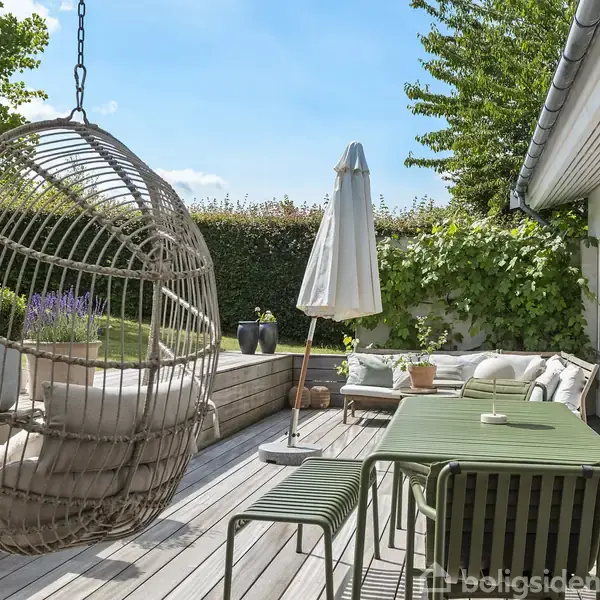 En hængekurvestol svinger på en terrasse med trædæk. Havebord med stole og parasol står nær en grøn sofa. Omgivet af planter og en hæk under en klar blå himmel.